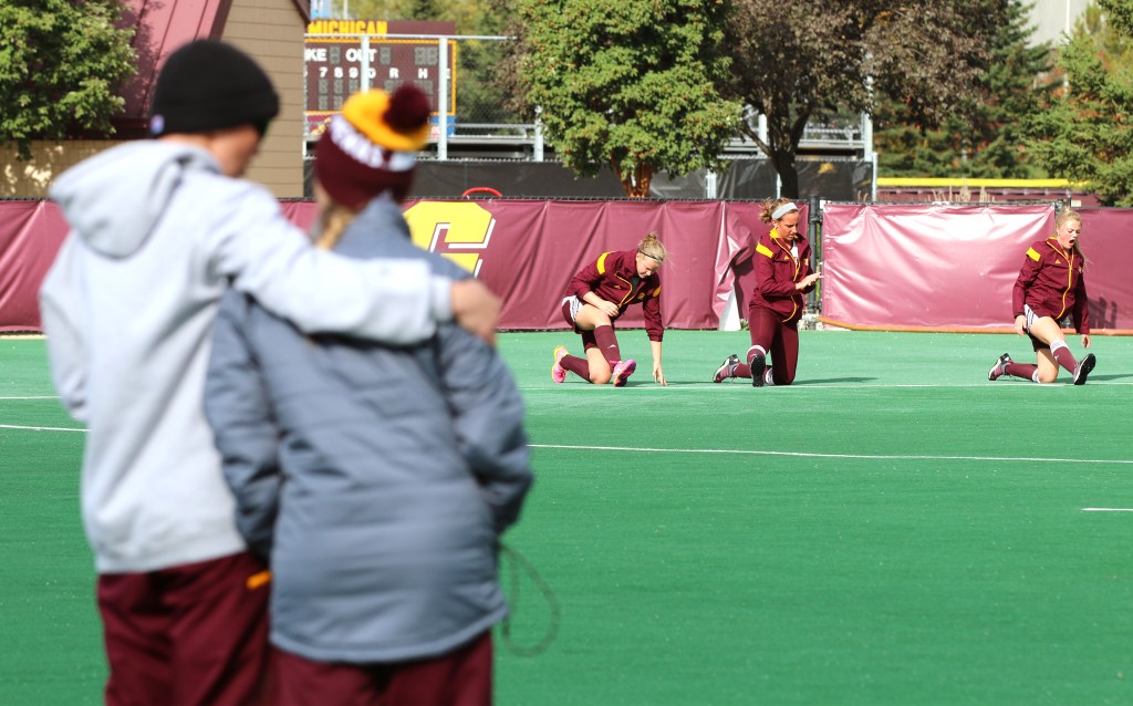 Maude Stalars, center, and teammates stretch while Head Coach Molly Pelowski and Assistant Coach Emily Girasole hug in the foreground at field hockey practice on Friday, Oct. 9, 2015. Before practice started, the head coach held a 